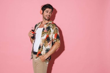 Pembe arka planda izole edilmiş gazoz içerken kablosuz kulaklık kullanan neşeli genç adamın portresi