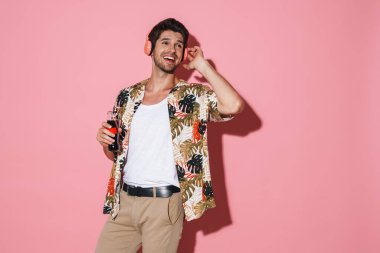 Pembe arka planda izole edilmiş gazoz içerken kablosuz kulaklık kullanan neşeli genç adamın portresi