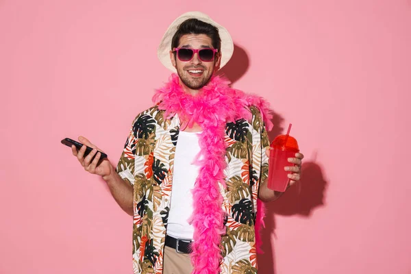 ピンクの背景に隔離されたソーダを飲みながら 携帯電話を使用してボアを身に着けている笑顔の男の肖像 — ストック写真