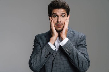 Tıraşsız, heyecanlı, ceketli iş adamının fotoğrafı. Gri duvarda izole edilmiş bir kamera karşısında şaşkınlığını ifade ediyor.