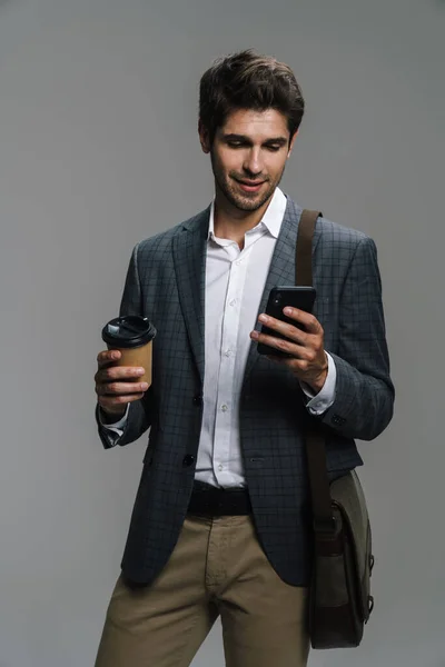 照片中带着微笑的自信商人一边喝着咖啡 一边用隔离在灰色墙壁上的手机 — 图库照片