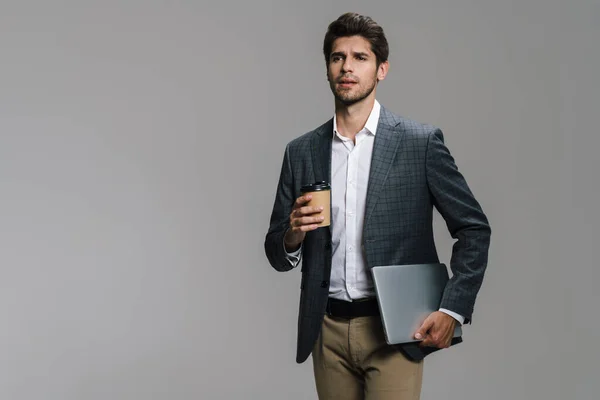 焦点を当てたUnshvenビジネスマンのコーヒーを飲むテイクアウトと灰色の壁に隔離されたノートパソコンを保持する写真 — ストック写真