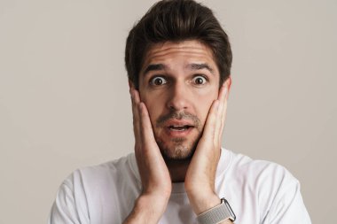 Temel tişörtlü, beyaz arka planda izole edilmiş kameraya şaşkınlığını ifade eden şok olmuş genç bir adamın portresi