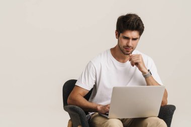 Tıraşsız, kafası karışık bir adamın portresi dizüstü bilgisayarla çalışırken beyaz arka planda izole bir şekilde koltukta oturuyor.