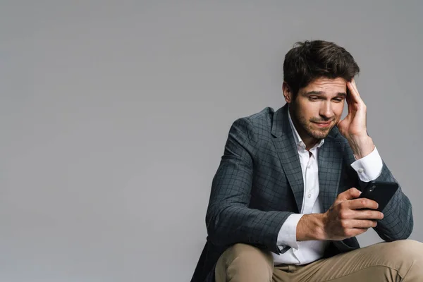 Photo Respectable Confused Businessman Using Cellphone While Siting Isolated Grey — Stock Photo, Image