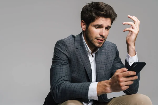 Photo Respectable Confused Businessman Using Cellphone While Siting Isolated Grey — Stock Photo, Image