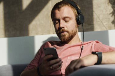 Evdeki kanepede otururken sakallı, cep telefonu ve kulaklık kullanan kızıl saçlı adamın fotoğrafı.