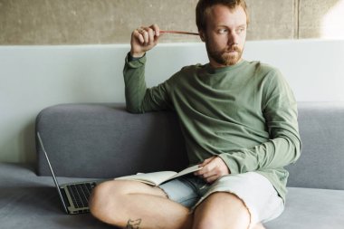 Evdeki kanepede otururken bilgisayarla çalışan ve notlar yazan kızıl saçlı adamın fotoğrafı.