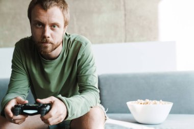 Odaklanmış kızıl saçlı adamın joystick ile oyun oynayıp evde patlamış mısır yediği fotoğraf.