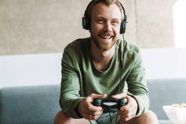 Evde kanepede otururken kulaklık takan ve joystick ile video oyunu oynayan gülen adamın fotoğrafı.