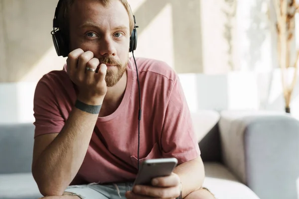 Zdjęcie Myślącego Rudzielca Brodą Pomocą Telefonu Komórkowego Słuchawek Podczas Siedzenia — Zdjęcie stockowe