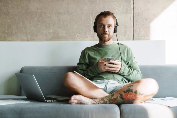 Foto Joven Satisfecho Con Uso Teléfono Móvil Auriculares Mientras Trabaja —  Fotos de Stock