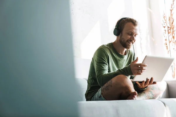 Foto Joven Complacido Sonriendo Mientras Usa Auriculares Computadora Portátil Sofá — Foto de Stock