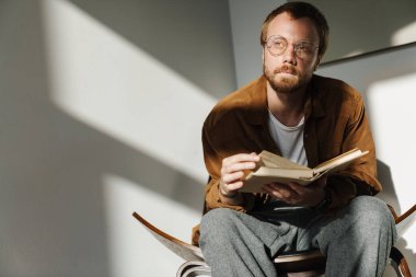 Evdeki koltukta otururken gözlüklü genç adamın kitap okuduğunu gösteren bir fotoğraf.