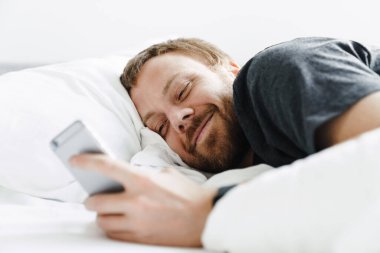 Gülümseyen sakallı adamın beyaz yatakta yatarken cep telefonu kullanmasının fotoğrafı.