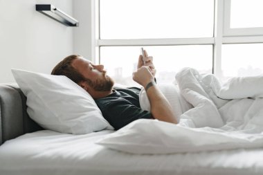 Odaklanmış sakallı adamın cep telefonu kullanırken beyaz yatakta yatarken çekilmiş fotoğrafı.