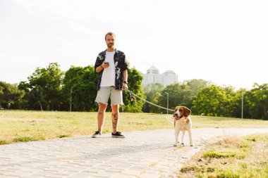 Kafkasyalı genç bir adamın yaz parkında av köpeğiyle yürürken cep telefonu kullanması.