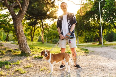 Beyaz sakallı genç bir adamın yaz parkında av köpeğiyle yürürken gülümsemesi.