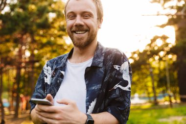 Yazlık parkta yürürken gülümseyen ve cep telefonu tutan beyaz sakallı genç bir adam.