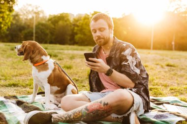 Kafkas sakallı genç bir adamın cep telefonu kullanması ve yaz parkında av köpeğiyle oturması.
