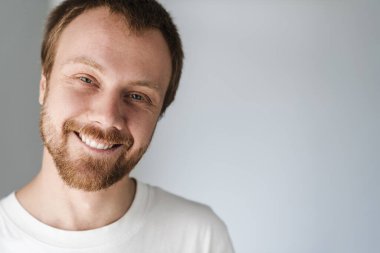 Gri arka planda izole edilmiş, kızıl saçlı, poz veren ve gülümseyen neşeli genç adamın fotoğrafı.