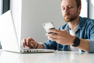 Kırmızı sakallı, cep telefonu ve dizüstü bilgisayar kullanan yakışıklı bir adamın fotoğrafı.