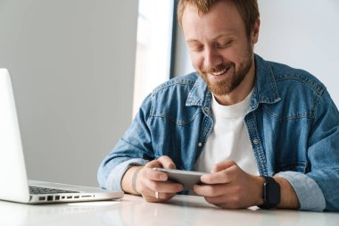 İçerideki masada dizüstü bilgisayarla çalışırken çevrimiçi oyun oynayan gülümseyen yakışıklı adamın fotoğrafı.
