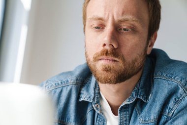 Photo of brooding handsome man wearing denim shit working with laptop while sitting at table indoors