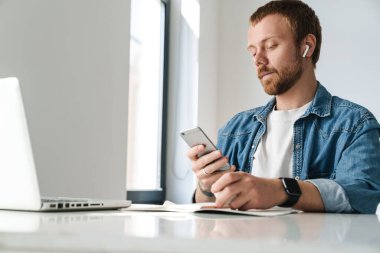 İçerideki masada dizüstü bilgisayarla çalışırken kablosuz kulaklık ve cep telefonu kullanan bir adamın fotoğrafı.