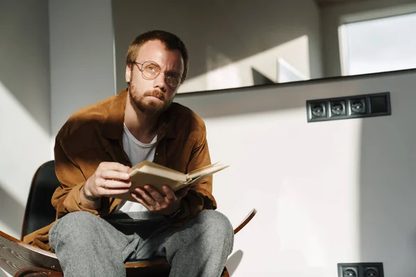 Foto Joven Pensante Con Anteojos Leyendo Libro Mientras Está Sentado —  Fotos de Stock