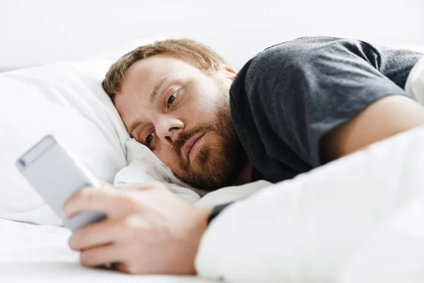 明るい部屋の白いベッドで寝た後に横たわっている間 携帯電話を使用して若い髭の男の写真 — ストック写真