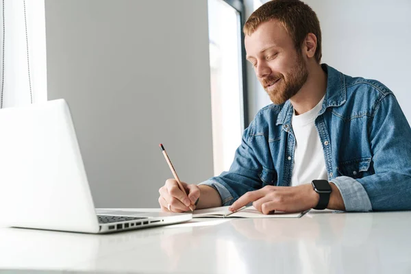 Fénykép Mosolygós Jóképű Férfi Írja Jegyzetek Miközben Dolgozik Laptop Asztalnál — Stock Fotó