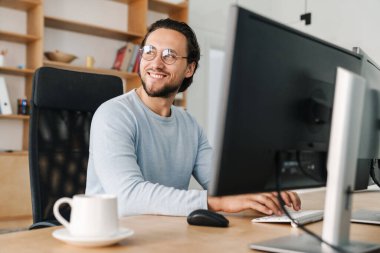 Ofiste bilgisayarla çalışan, gözlüklü, gülümseyen, traşsız programcı.