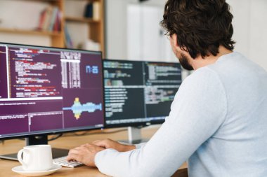 Ofisteki bilgisayarlarla çalışan, gözlüklü, traşsız programcının kesilmiş görüntüsü.