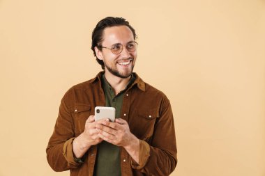 Gözlüklü, gülümseyen ve bej arka planda izole edilmiş cep telefonu kullanan heyecanlı kıllı adamın görüntüsü