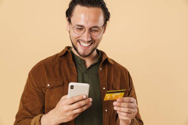 Kredi kartı tutan ve bej arka planda izole edilmiş cep telefonu kullanan neşeli kıllı adamın görüntüsü