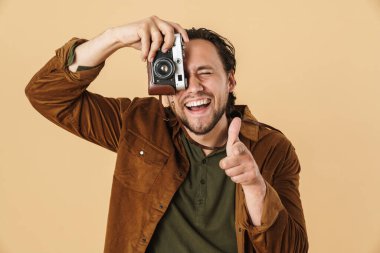 Gülen beyaz bir adamın retro kamerada fotoğraf çekmesi ve bej arka planda izole edilmiş bir parmak göstermesi.