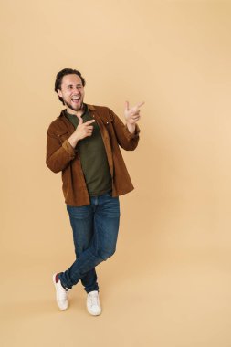 Yakışıklı, neşeli bir adamın bej arka planda izole edilmiş bir şekilde göz kırparak ve parmaklarını işaret etmesi.