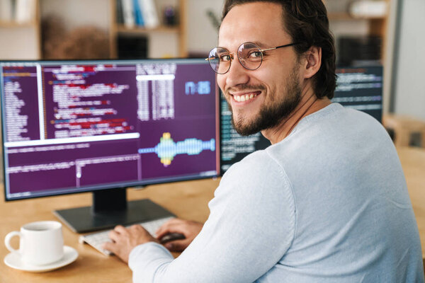 Image of smiling unshaven programmer man wearing eyeglasses working with computers in office