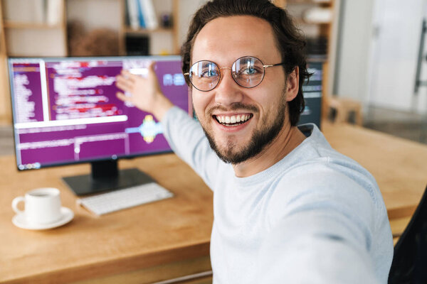 Image of unshaven excited programmer man showing screens while taking selfie photo in office