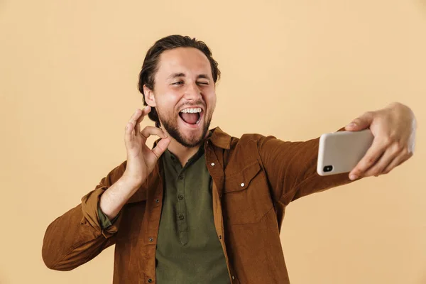 照片中快乐的男人在手机上用米色背景的手机自拍时显示出了好的签名 — 图库照片