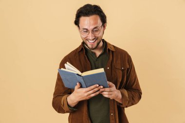 Gözlüklü neşeli, tıraşsız bir adamın resmi kitap okuyor ve bej arka planda izole bir şekilde gülümsüyor.