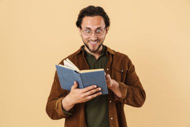 Gözlüklü heyecanlı, tıraşsız bir adamın resmi kitap okuyor ve bej arka planda izole bir şekilde gülümsüyor.