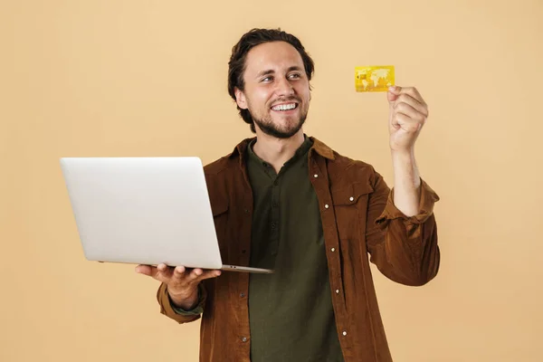 照片中快乐的未剃光头发的男人拿着信用卡 使用的笔记本电脑与米色背景相分离 — 图库照片