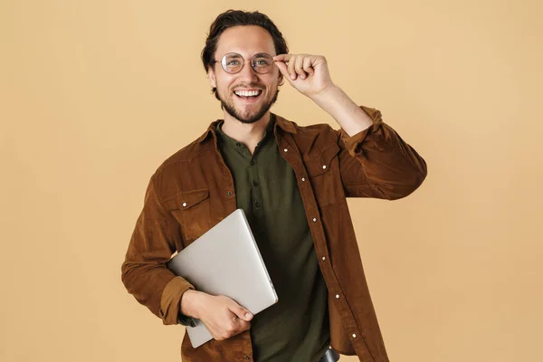 图片中 穿着眼镜的快乐的未剃光的男人在使用笔记本电脑时 在米色背景下露出微笑 — 图库照片