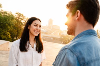 Açık havada yürürken gülümseyen ve birbirlerine bakan romantik çok kültürlü çiftin görüntüsü.