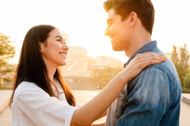 Romantik, çok kültürlü bir çiftin açık havada kucaklaşırken gülümseyip birbirlerine bakması.