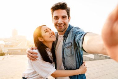 Gülümseyen çok kültürlü bir çiftin dışarıda yürürken kucaklaşması ve selfie çekmesi.