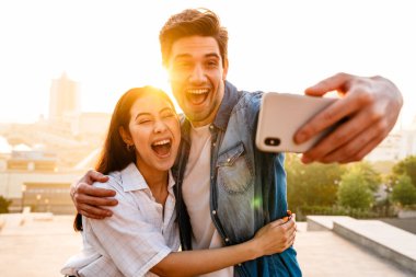 Heyecanlı, çok kültürlü bir çiftin cep telefonuyla kucaklaşıp selfie çektiği bir görüntü.