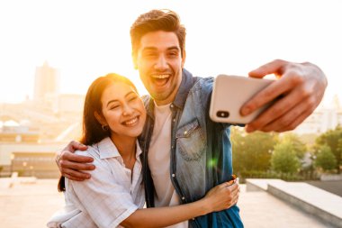Heyecanlı, çok kültürlü bir çiftin cep telefonuyla kucaklaşıp selfie çektiği bir görüntü.